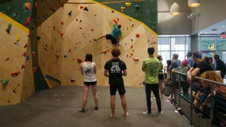 three students watch another student climb the rock wall