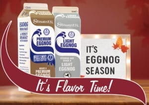 A promotional display featuring Stewart's Shops eggnog products. Three cartons are shown: Premium Eggnog, Light Eggnog, and a carton displaying an eggnog French toast recipe. A decorative sign in the background reads 'It's Eggnog Season' with autumn leaves attached to the corner. The scene is set against a warm, autumnal background with falling leaves. A maroon banner with white text reading 'It's Flavor Time!' overlays the bottom of the image.
