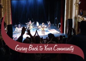 An audience applauds a live musical performance on stage in an ornate theater, with a banner overlay that reads 'Giving Back to Your Community.