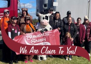 A group of Stewart's Shops employees and mascots pose outdoors in front of a large truck, holding a red Stewart's Shops banner. The group includes people in maroon uniforms, casual clothing, and two individuals dressed in bear costumes. A person in a cow mascot costume stands in the center, holding the banner. A young child in a maroon jacket stands in front of the group. A maroon banner with white text reading 'We Are Closer To You!' overlays the bottom of the image.