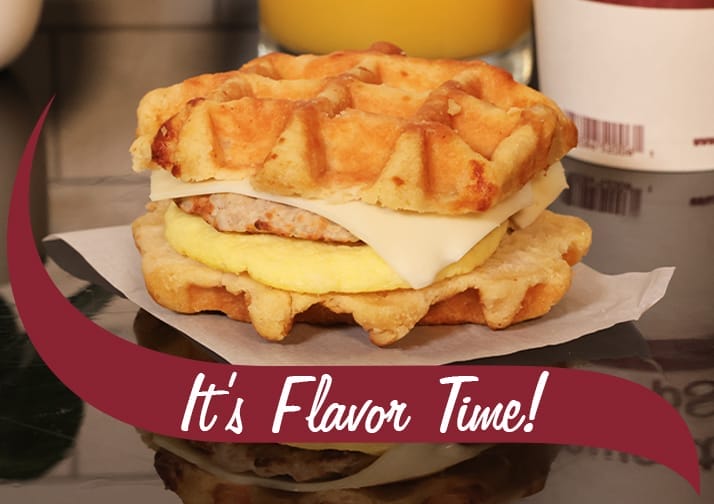 A close-up of a breakfast sandwich made with two golden brown waffles as the bun, containing a sausage patty, egg, and a slice of melted cheese. The sandwich is placed on a piece of parchment paper on a reflective surface. In the background, a cup of coffee and a glass of orange juice are partially visible. A maroon banner with white text reading 'It's Flavor Time!' overlays the bottom of the image.