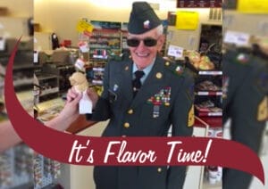An elderly veteran wearing a decorated military uniform and sunglasses smiles while receiving an ice cream cone inside a Stewart's Shops store. Shelves stocked with various products are visible in the background. A maroon banner with white text reading 'It's Flavor Time!' overlays the bottom of the image.