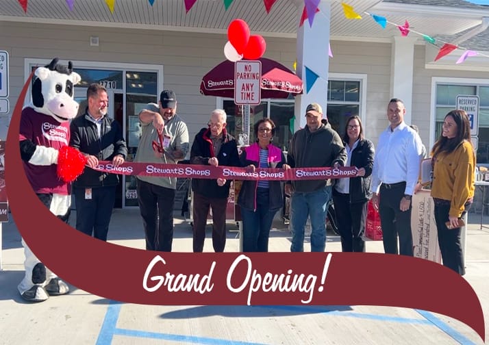 A group of people gathers in front of a Stewart's Shops store for a grand opening ribbon-cutting ceremony. A person in a cow mascot costume stands on the left holding the ribbon, while a man in a cap cuts it with large scissors. Several individuals, including store employees and community members, stand behind the ribbon, smiling. The store's entrance is decorated with colorful pennant banners and red and white balloons. A maroon banner with white text reading 'Grand Opening!' overlays the bottom of the image.