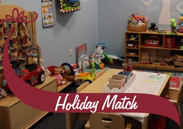 A colorful children's playroom filled with toys, books, and activities. A small table with chairs is set up with coloring sheets and art supplies. Shelves in the background hold wooden toys, stuffed animals, and baskets of supplies. A red toy car and a plush doll are positioned near a wooden crib. The walls are painted light blue and decorated with educational posters and artwork. A maroon banner with white text reading 'Holiday Match' overlays the bottom of the image, with a decorative bow graphic on the left side.