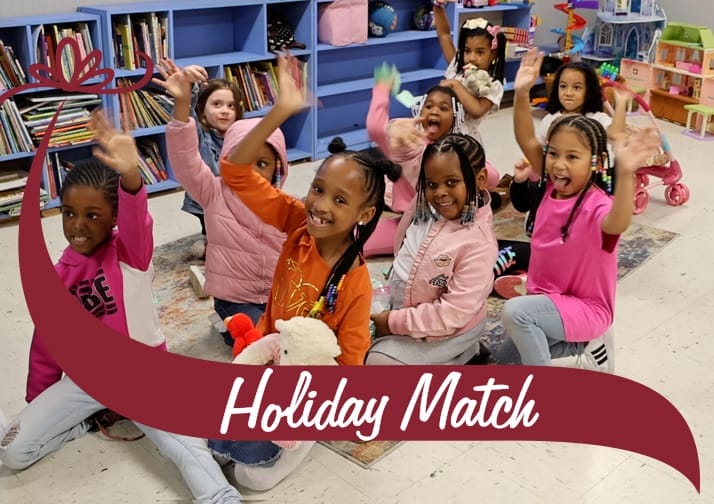 A group of young children sitting on a colorful rug in a classroom, smiling and waving at the camera. They are dressed in bright clothing and holding stuffed animals. Behind them are blue bookshelves filled with books and various toys. A maroon banner with white text reading 'Holiday Match' overlays the bottom of the image, with a decorative bow graphic on the left side.