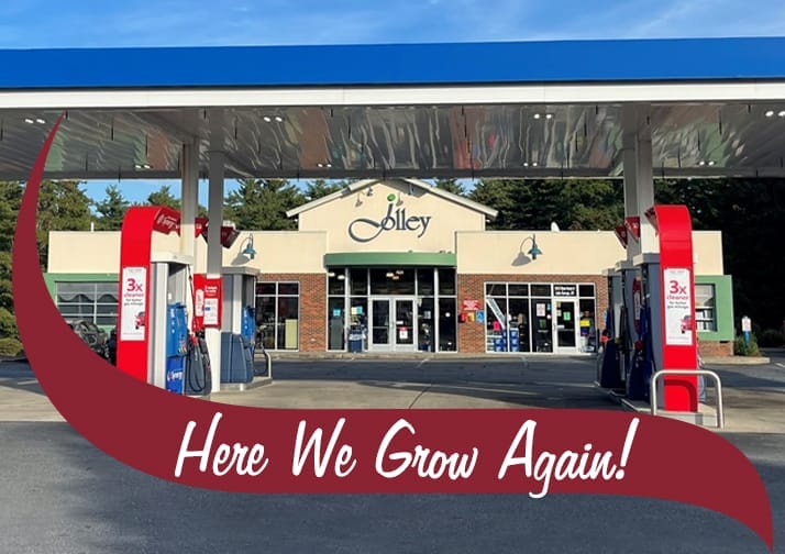 A Jolley convenience store and gas station with a blue and white canopy over multiple fuel pumps. The store has a beige and brick exterior with large glass windows and doors. The Jolley logo is displayed above the entrance, and promotional signage is visible on the gas pumps. The background features green trees and a clear blue sky. A maroon banner with white text reading 'Here We Grow Again!' overlays the bottom of the image.