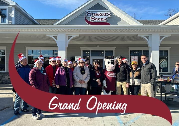 A group of people wearing festive Santa hats stand in front of a newly opened Stewart's Shops store, posing for a group photo. The store has a gray exterior with white trim and a red Stewart's Shops sign above the entrance. A person in a cow mascot costume wearing a maroon Stewart's Shops shirt stands in the middle of the group. The event is taking place on a sunny day with clear blue skies. A maroon banner with white text reading 'Grand Opening' overlays the bottom of the image.