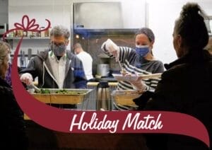Two volunteers wearing face masks and gloves serve food to people at a community kitchen. One volunteer, wearing a striped sweater, is scooping food onto a plate, while the other, in a black jacket, is preparing salad trays. A maroon banner with white text reading 'Holiday Match' overlays the bottom of the image, with a decorative bow graphic on the left side. The background features a stainless steel kitchen with cooking equipment and utensils.