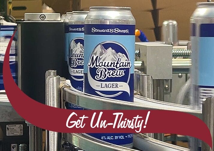 Conveyor belt carrying cans of Stewart's Shops Mountain Brew Lager. The cans have a blue and white label featuring an illustration of mountains and the brand name in bold script. A maroon banner with white text reading 'Get Un-Thirsty!' overlays the bottom right of the image. The background shows industrial equipment and stacked boxes, indicating a production setting.