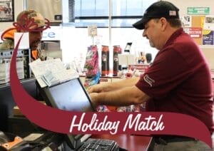 A store employee in a maroon uniform and black cap is assisting a customer at the checkout counter. The customer, wearing a camouflage jacket and a baseball cap, is engaged in conversation with the employee. The counter is filled with receipts, a cash register, and various products, including snacks and beverages. A maroon banner with white text reading 'Holiday Match' overlays the bottom of the image, with a decorative bow graphic on the left side. Behind the counter, shelves are stocked with coffee and other convenience store items, with a bright window in the background.