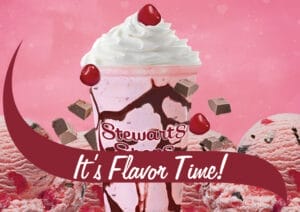 A pink milkshake topped with whipped cream and a cherry in a Stewart's Shops cup, drizzled with chocolate syrup. Surrounding the shake are scoops of pink ice cream with chocolate chunks and cherries, with chocolate pieces floating in the air. The background is a pink gradient with subtle heart shapes. A maroon banner with white text reading 'It's Flavor Time!' overlays the bottom of the image.