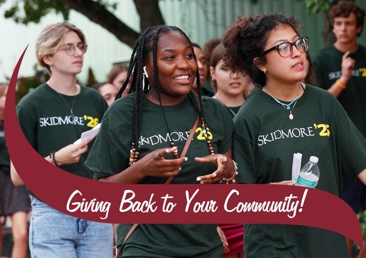 A group of young people wearing matching green t-shirts with 'Skidmore '23' printed on them walk together outdoors, smiling and engaged in conversation. Two young women in the foreground are holding papers and a water bottle. A maroon banner with white text reading 'Giving Back to Your Community!' overlays the bottom of the image. Trees and a building are visible in the background.