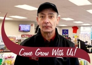 A male store manager wearing a black cap and black jacket with a name tag reading 'Patrick, Manager 8 Yrs' stands inside a convenience store. He has a serious expression and looks directly at the camera. The background features store shelves stocked with various products, a digital screen on the wall, and promotional signage. A maroon banner with white text reading 'Come Grow With Us!' overlays the bottom of the image.