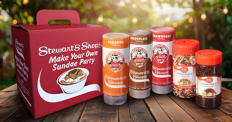 Stewart’s Shops 'Make Your Own Sundae Party' kit displayed on a wooden table with a blurred outdoor background. The kit includes a red box labeled 'Make Your Own Sundae Party,' three squeeze bottles of Stewart’s Shops toppings (caramel, chocolate, and strawberry), and two containers of Betty Crocker 'Parlor Perfect!' sprinkles (rainbow sprinkles and chocolate sprinkles).